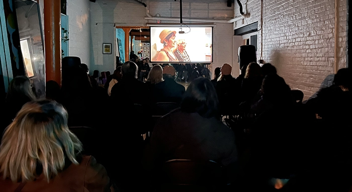 Audience seated in a dimly lit room watching Prodigal Daughter projected on a screen during an indoor community screening.